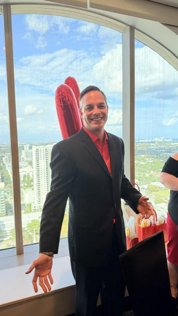 Man in black suit and red shirt holding red boxing gloves by high-rise window overlooking city