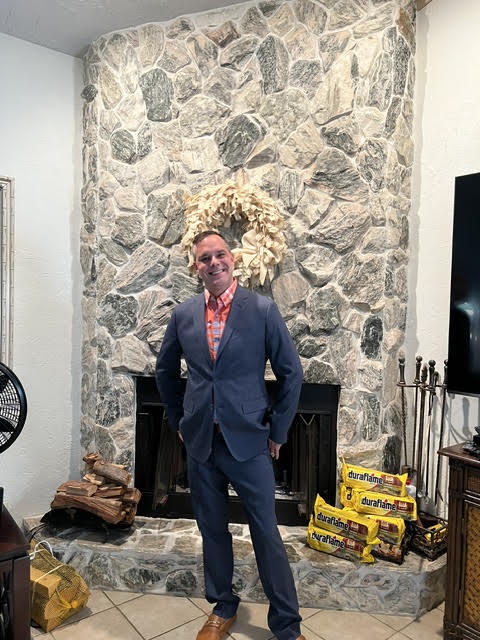 Man in navy blazer and red shirt standing before stone fireplace with yellow equipment boxes