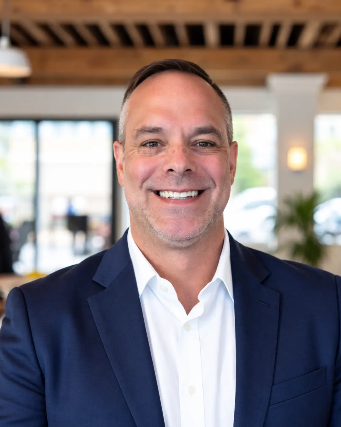 Professional headshot of man in navy blazer and white dress shirt smiling in modern office lobby