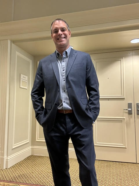 Professional man in navy blazer and light blue shirt standing in hallway with hands in pockets